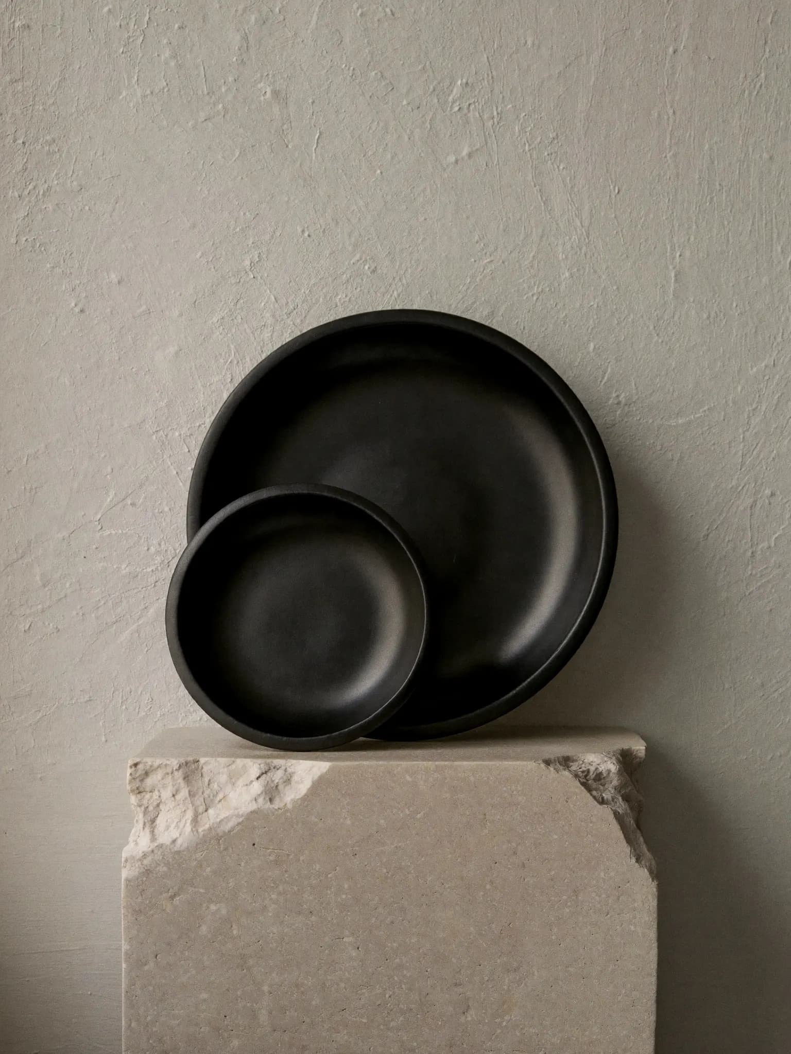 Deep Saucer, set of 2 stoneware piece in charcoal glaze, on a stone block against a plaster wall