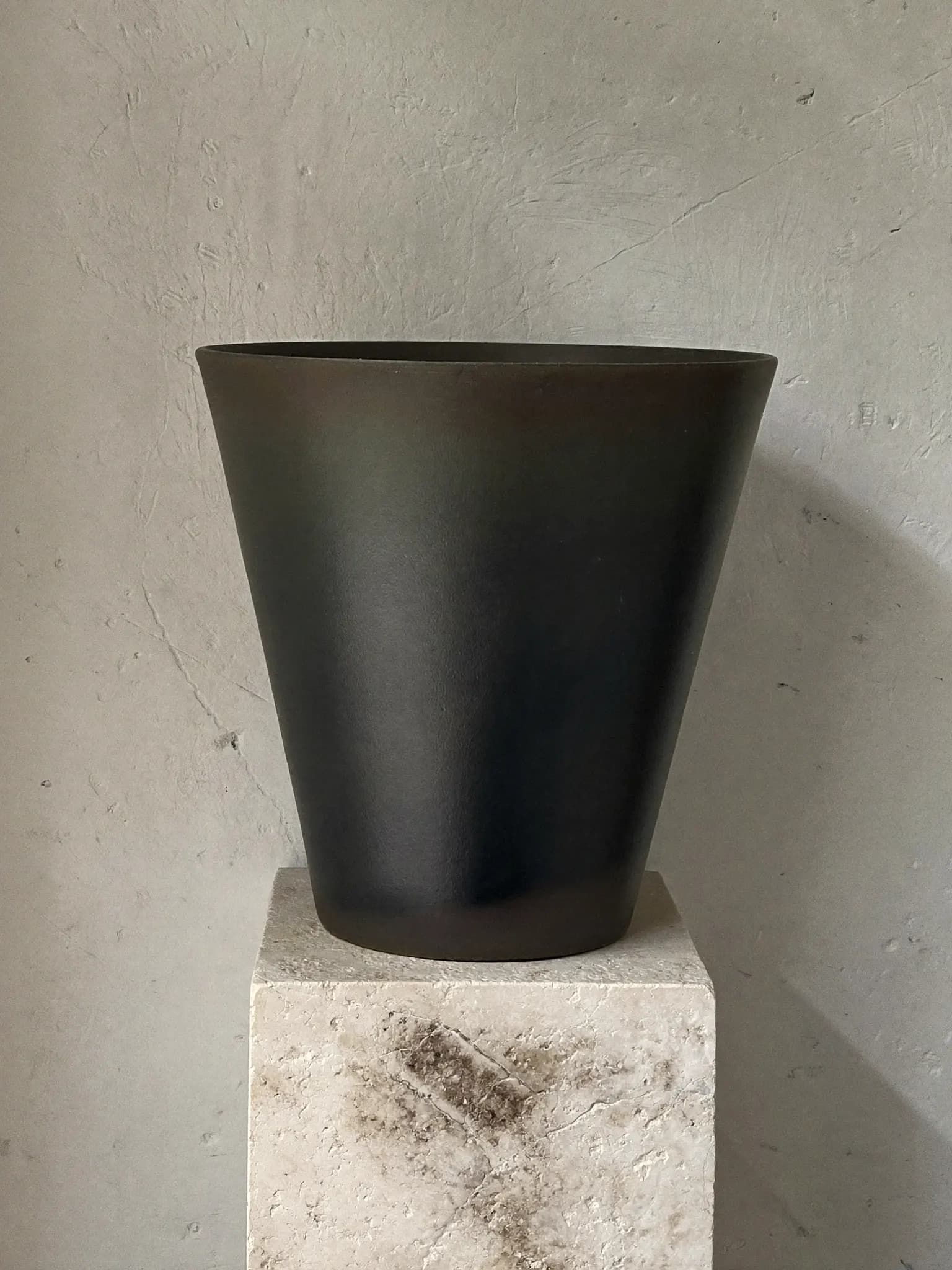 Taper stoneware planter in charcoal glaze, on a stone block against a plaster wall